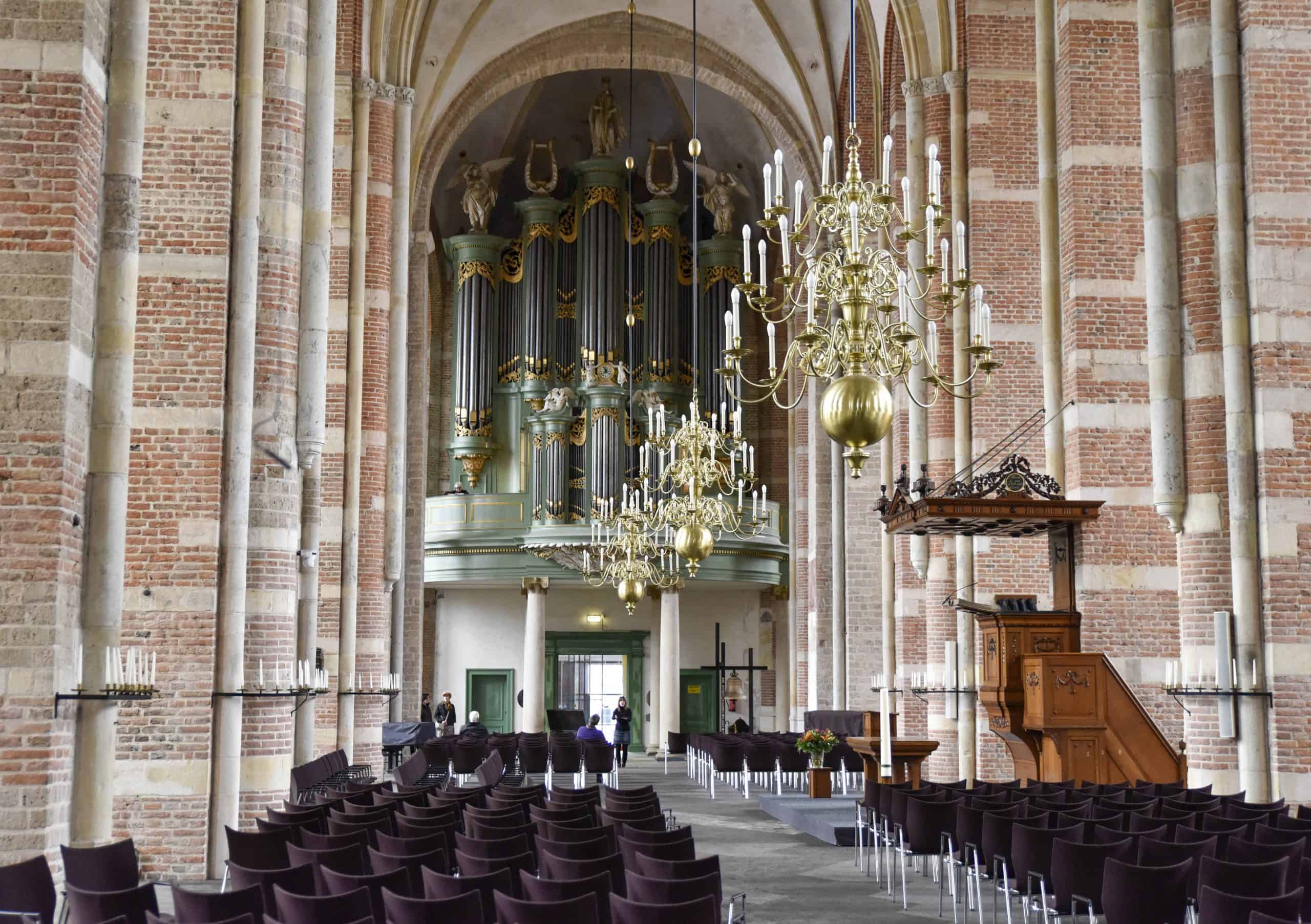 Interieur Grote Kerk Dordrecht met orgel, kroonluchters en rijen stoelen. Bakstenen pilaren en houten preekstoel.