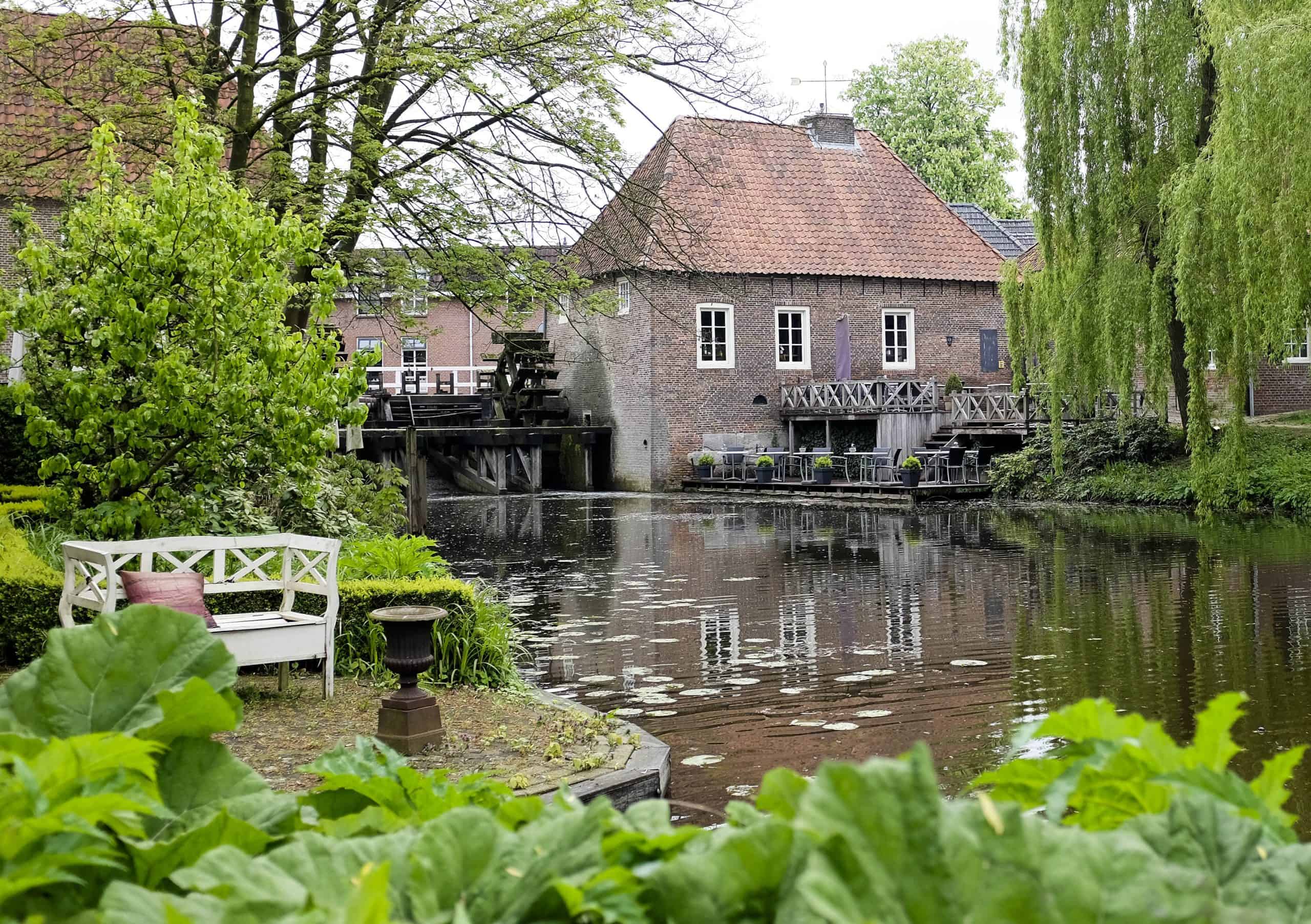 Historische watermolen met terras aan het water, omringd door groen in een rustige omgeving.