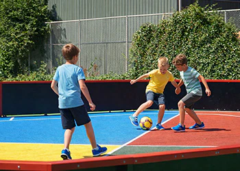 Jongens spelen voetbal op een kleurrijk, klein voetbalveld met groene begroeiing op de achtergrond.