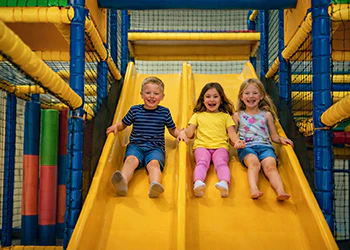 Drie vrolijke kinderen glijden van een gele glijbaan in een binnenspeeltuin.
